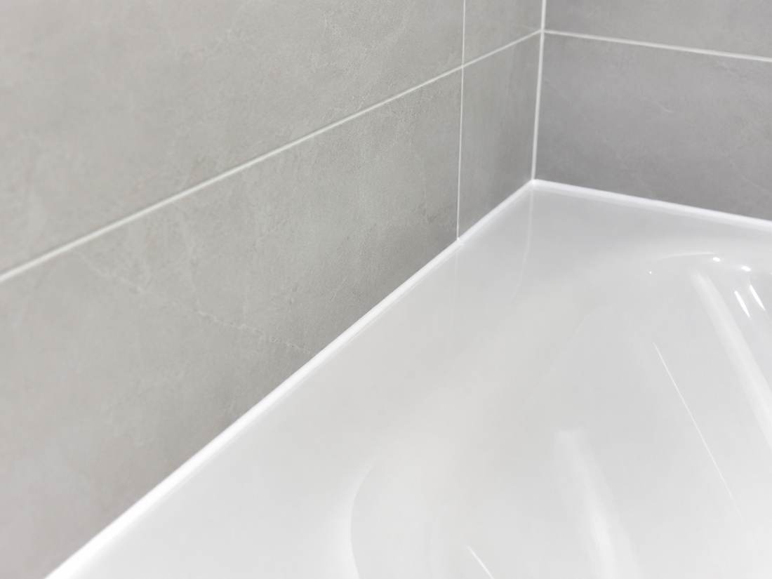 Close-up of a resealed bathtub edge with clean, new white sanitary sealant against grey tiling